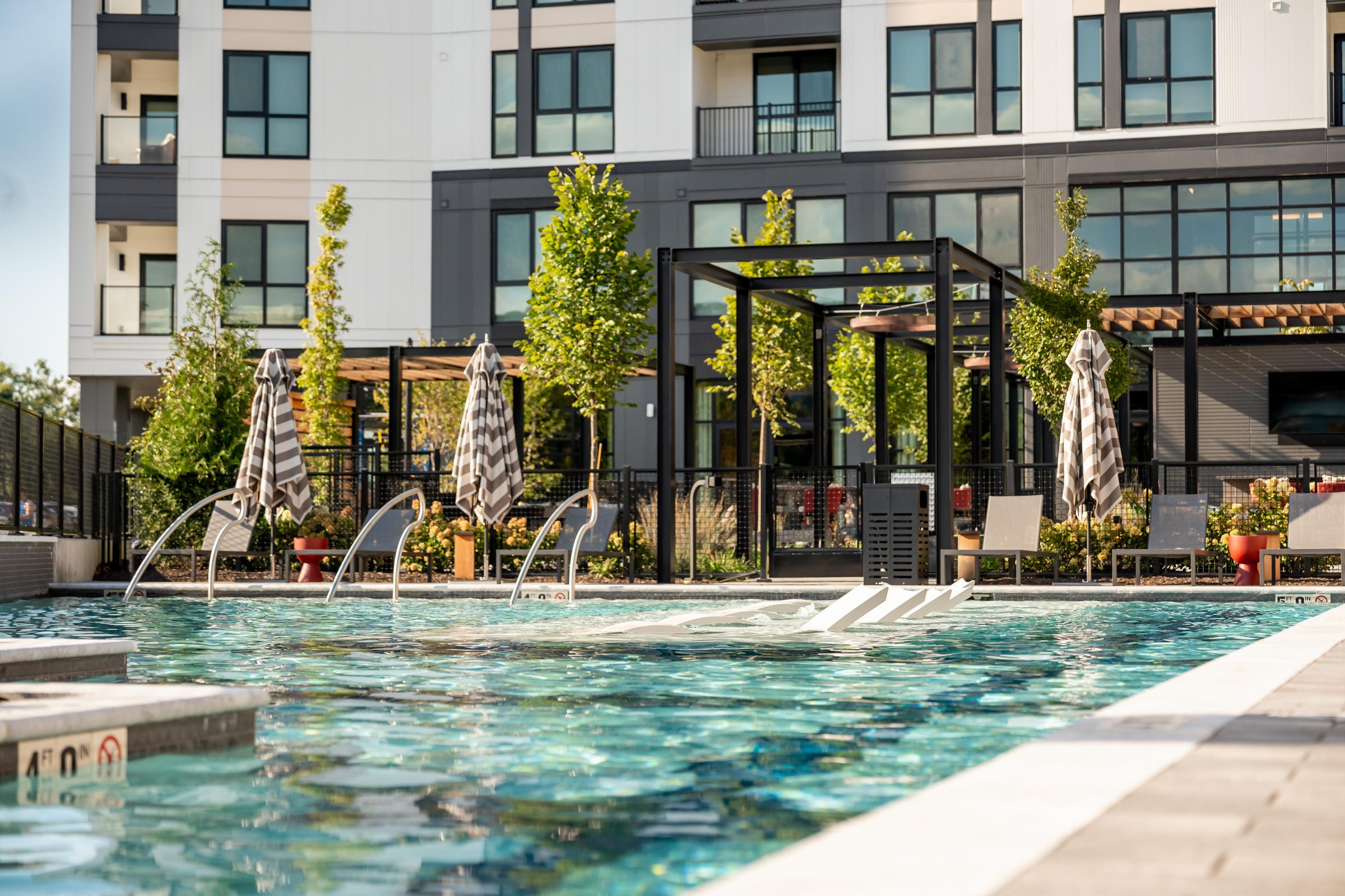 Madison West Elm apartments Modern apartment building with a pool, lounge chairs, umbrellas, and green plants on a sunny day.