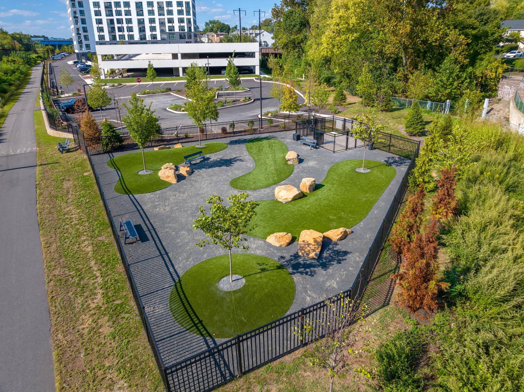 Madison West Elm apartments Aerial view of a fenced dog park with benches, trees, rocks, and winding paths near a parking lot.