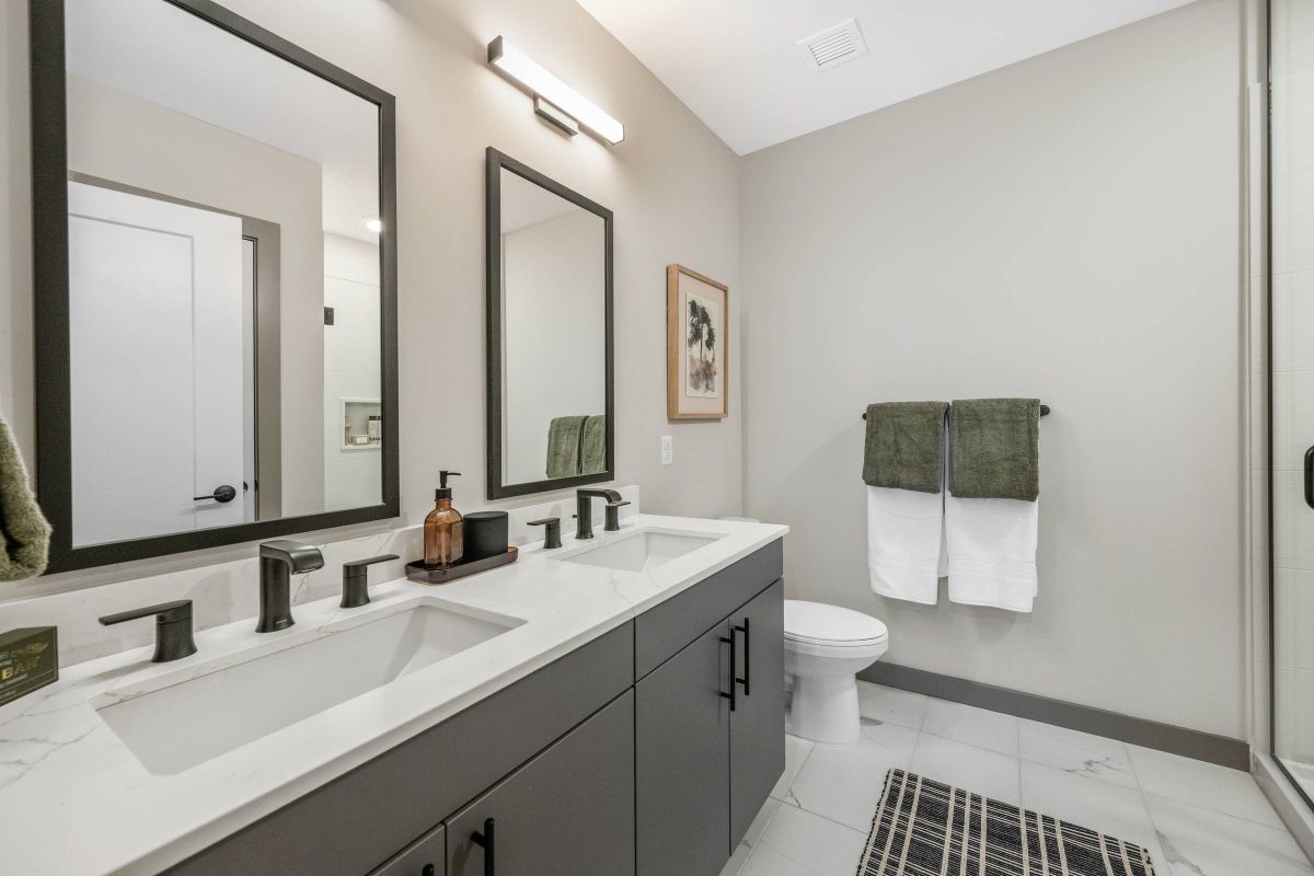 Madison West Elm apartments Modern bathroom with double sinks, two mirrors, towels, and a rug on a light tile floor.